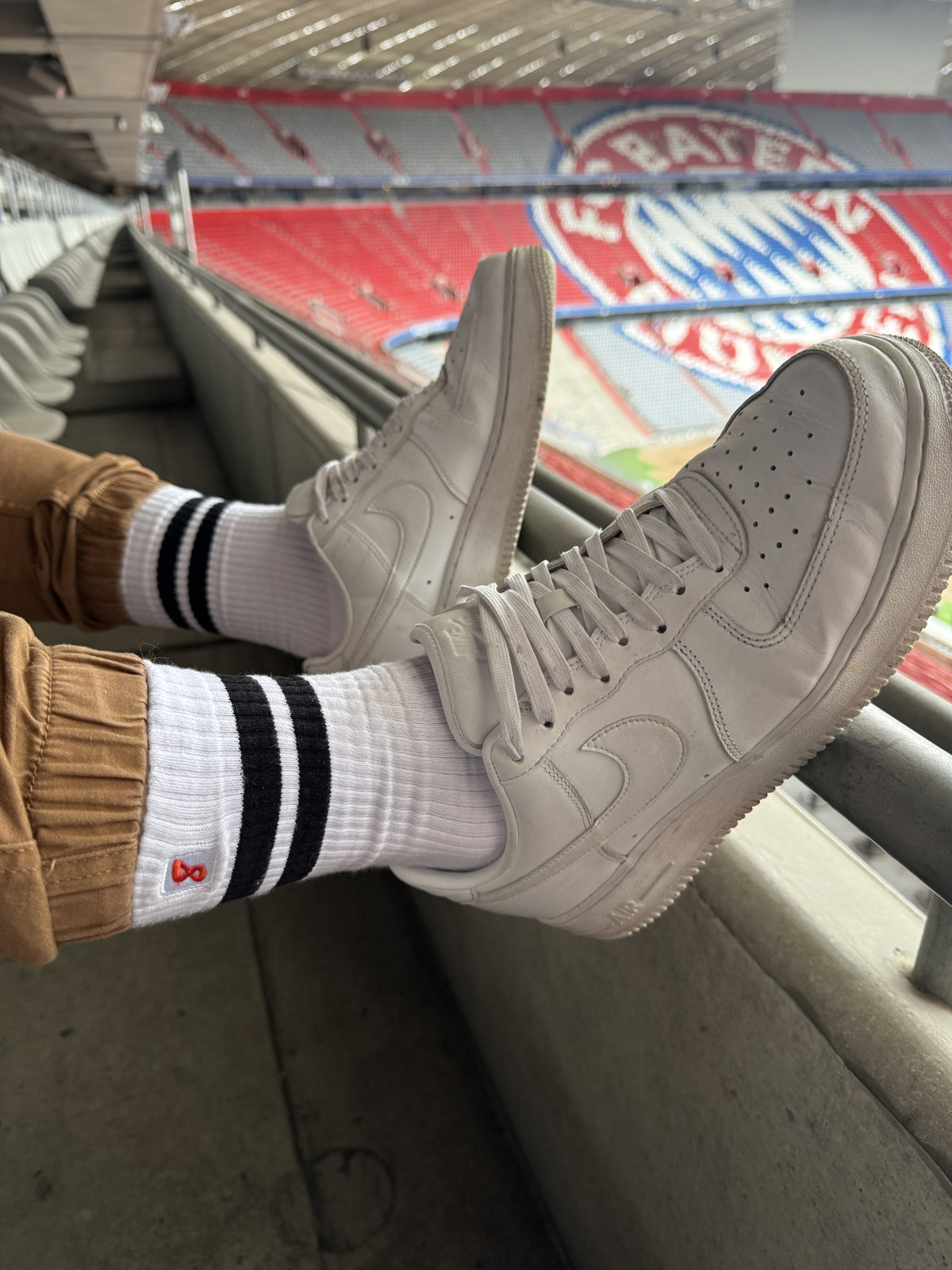 Gros plan sur des chaussettes rétro Sk8erboy® à rayures noires, portées avec des baskets Nike blanches au stade Allianz Arena de Munich, look gay sportif FC Bayern à fort impact.