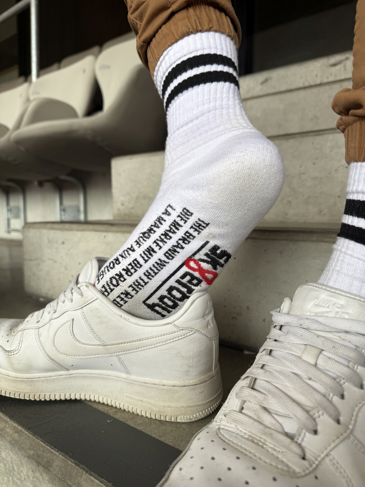 Moment de stade décontracté : chaussettes rétro Sk8erboy® à rayures noires et imprimé voyant sous la semelle, associées à des Nike Air Force 1 blanches – un look streetwear stylé avec attitude.
