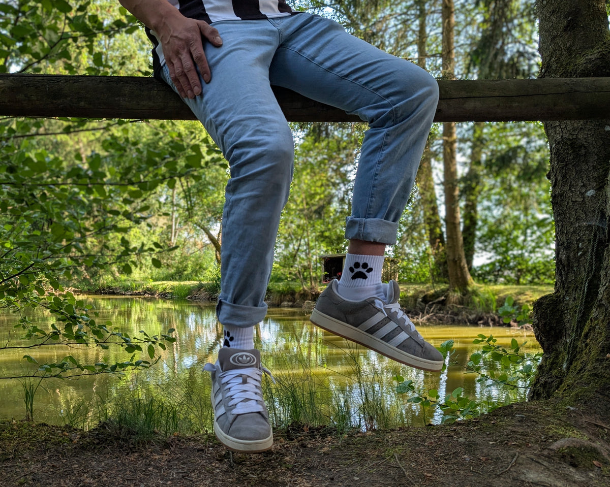 Man sitzt in der Natur auf einem Holzbalken, trägt hochgekrempelte Jeans, graue Sneaker und weiße Sk8erboy® Socks mit schwarzem Pawprint-Motiv – entspannter Outdoor-Vibe mit Fetisch-Flair.