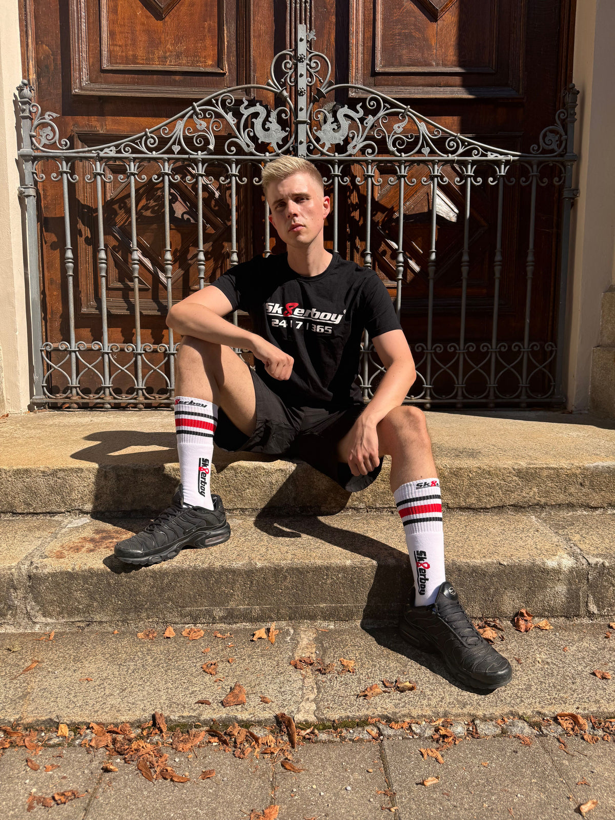 hübscher gay twink trägt ein sk8erboy t-shirt shirt in schwarz baumwolle cotton mit einem sk8erboy aufdruck mit weisser schrift und 8 acht in rot mit 24 7 365 fetter aufdruck
