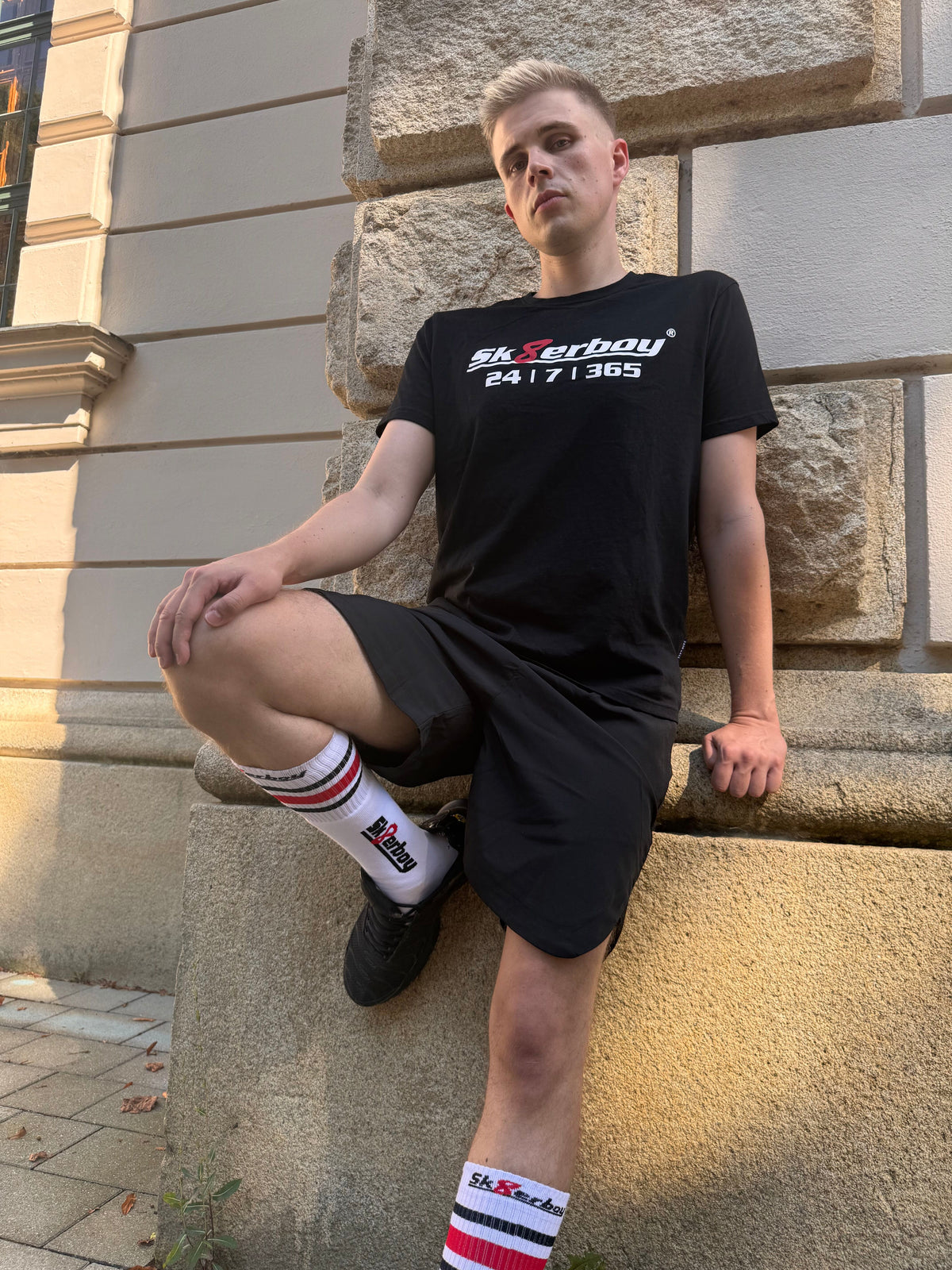 hübscher gay twink trägt ein sk8erboy t-shirt shirt in schwarz baumwolle cotton mit einem sk8erboy aufdruck mit weisser schrift und 8 acht in rot mit 24 7 365 fetter aufdruck