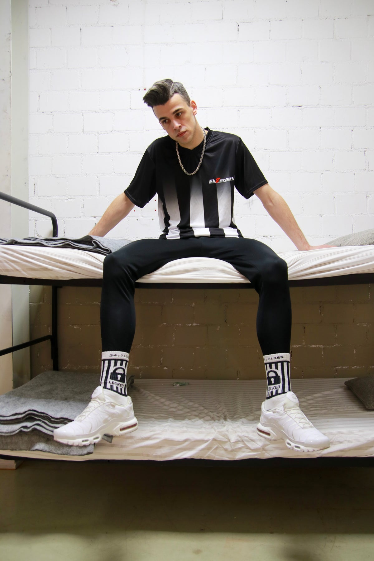 Urbaner Look: Model sitzt mit dem Sk8erboy® Tikot und glänzender Jogginghose in einem Stockbett in einer Gefängniszelle