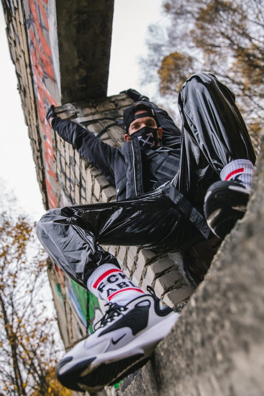 cooler gay mit pvc sporthose streckt sich an einer steinwand mit fck abl socks und nike air max sneaker im bild