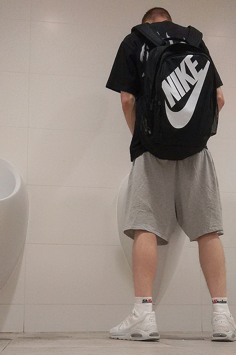 young gay man in short sports clothes wearing a Nike backpack standing in front of a urinal, with Nike sneakers and white sk8erboy quarter socks