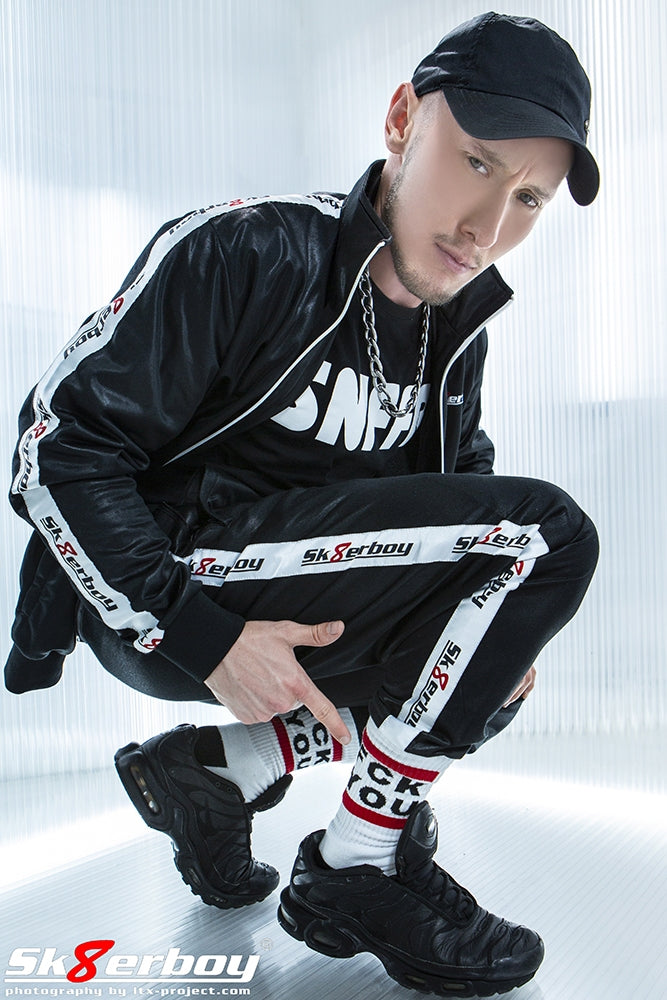 junger gay mit sportlichem glanz trainingsanzug von sk8erboy und snffr t-shirt in schwarz zeigt auf seine fck you socks von sk8erboy in schwarzen nike tn sneaker