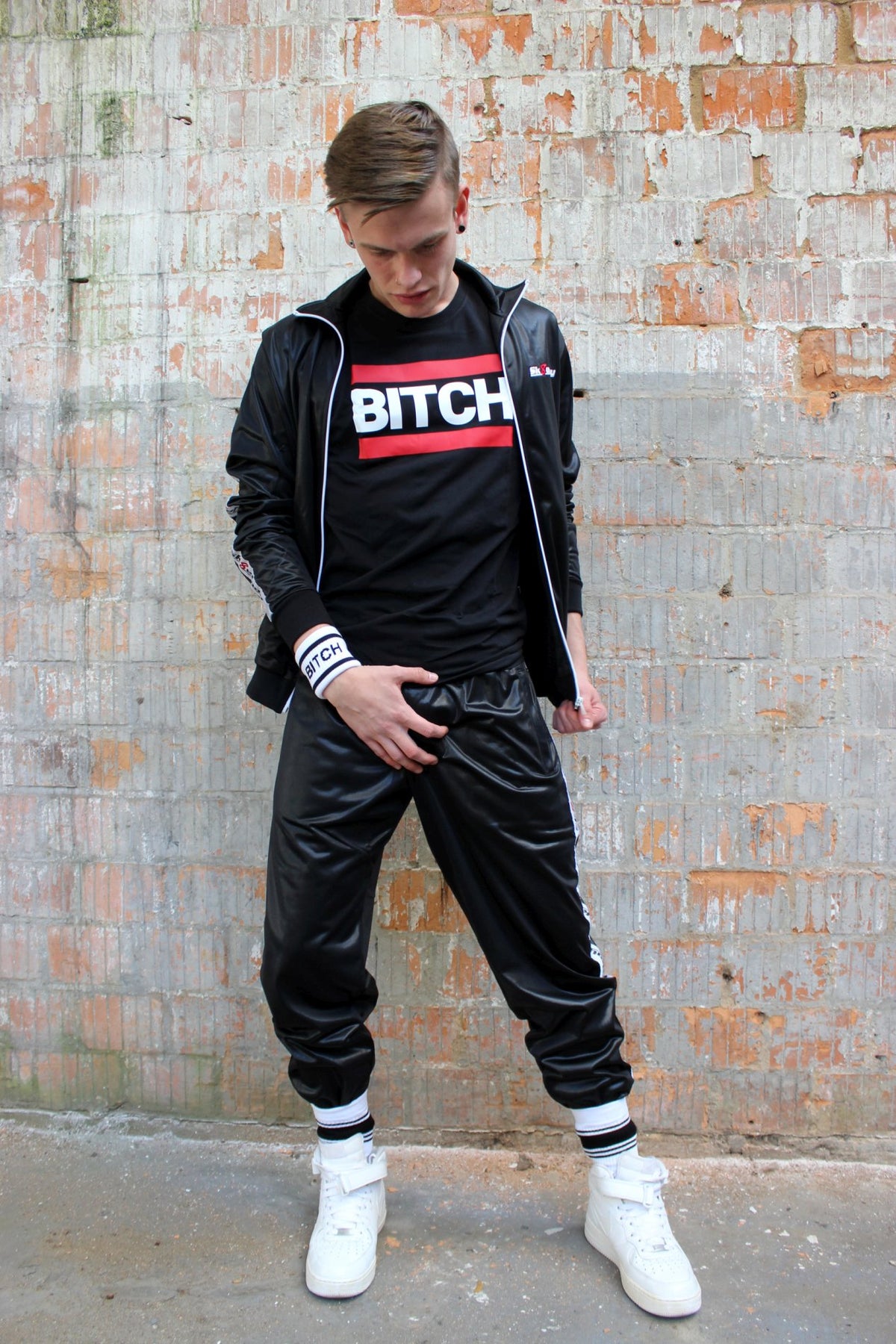 Boy in a shiny sk8erboy tracksuit wears a black T-shirt with a bitch print and holds his crotch, looking at his white Nike sneakers and socks by Nike and sk8erboy.