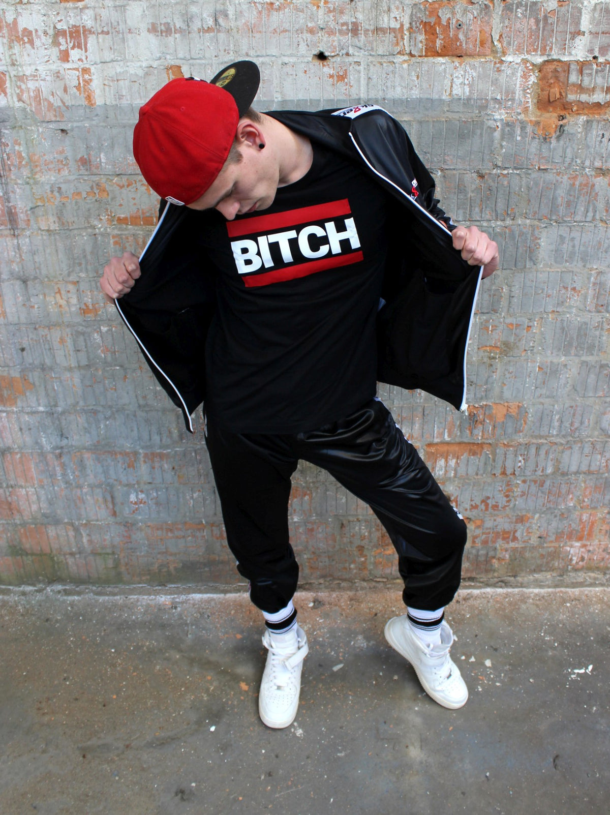 Young man in front of a wall wearing an open shiny jacket made of glossy fabric, with a black T-shirt reading BITCH in white capital letters and red bars above and below