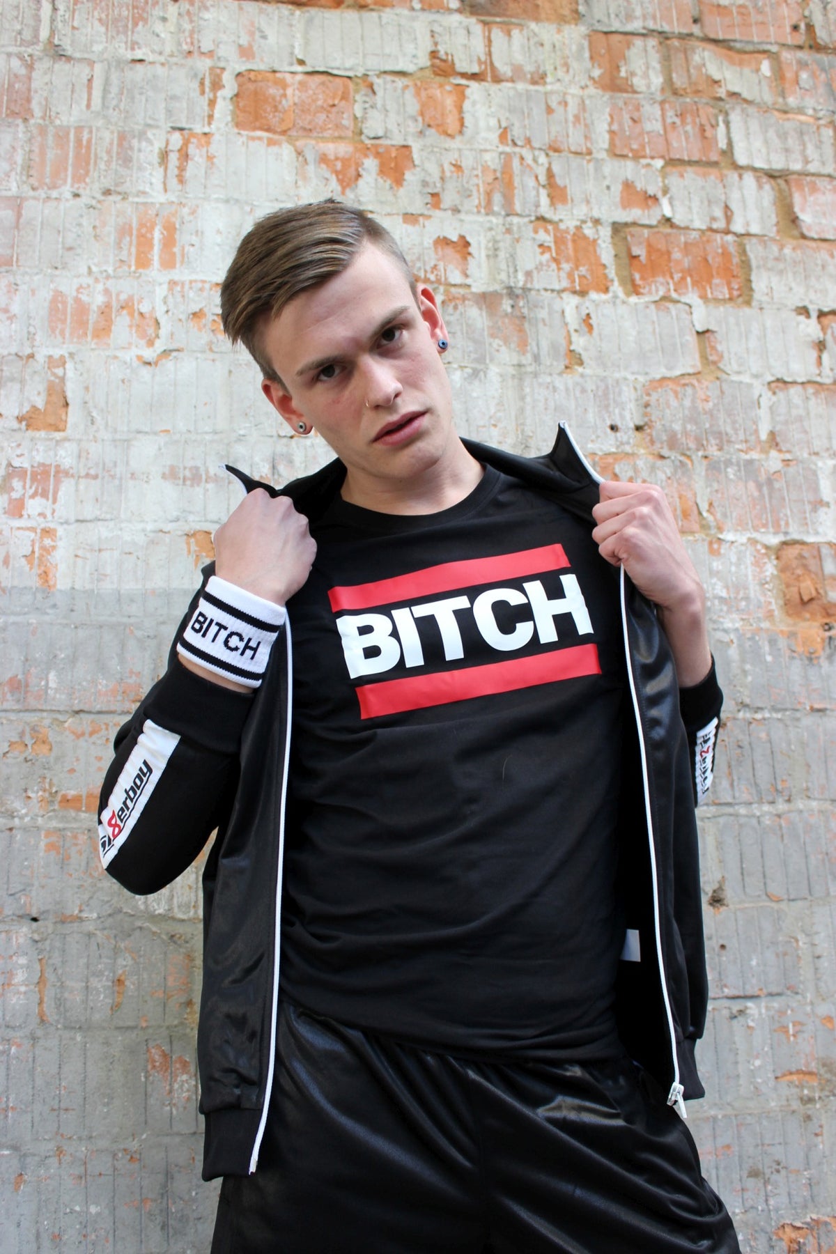 Young gay man in an open shiny tracksuit jacket showing a black T-shirt printed with "bitch" in white, with red bars above and below.