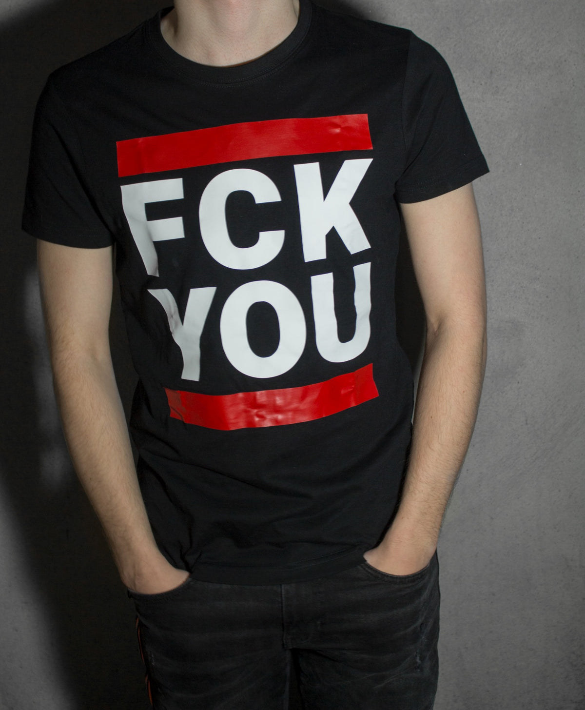 Boy in front of a wall wearing a black sk8erboy T-shirt with the print "fck you" in large white letters with red bars