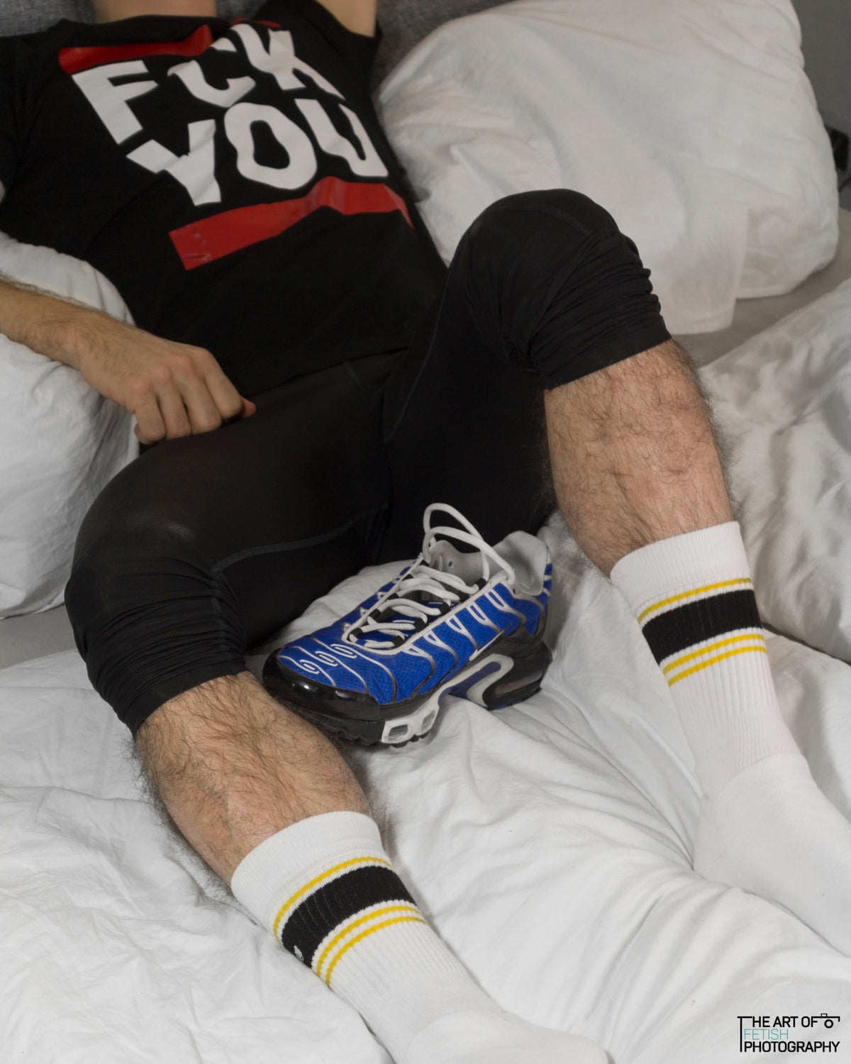 Young man lying in bed wearing sweatpants and Nike TN sneakers with yellow Sk8erboy Deluxe socks, wearing a black T-shirt with the print "fck you" in white with red bars.