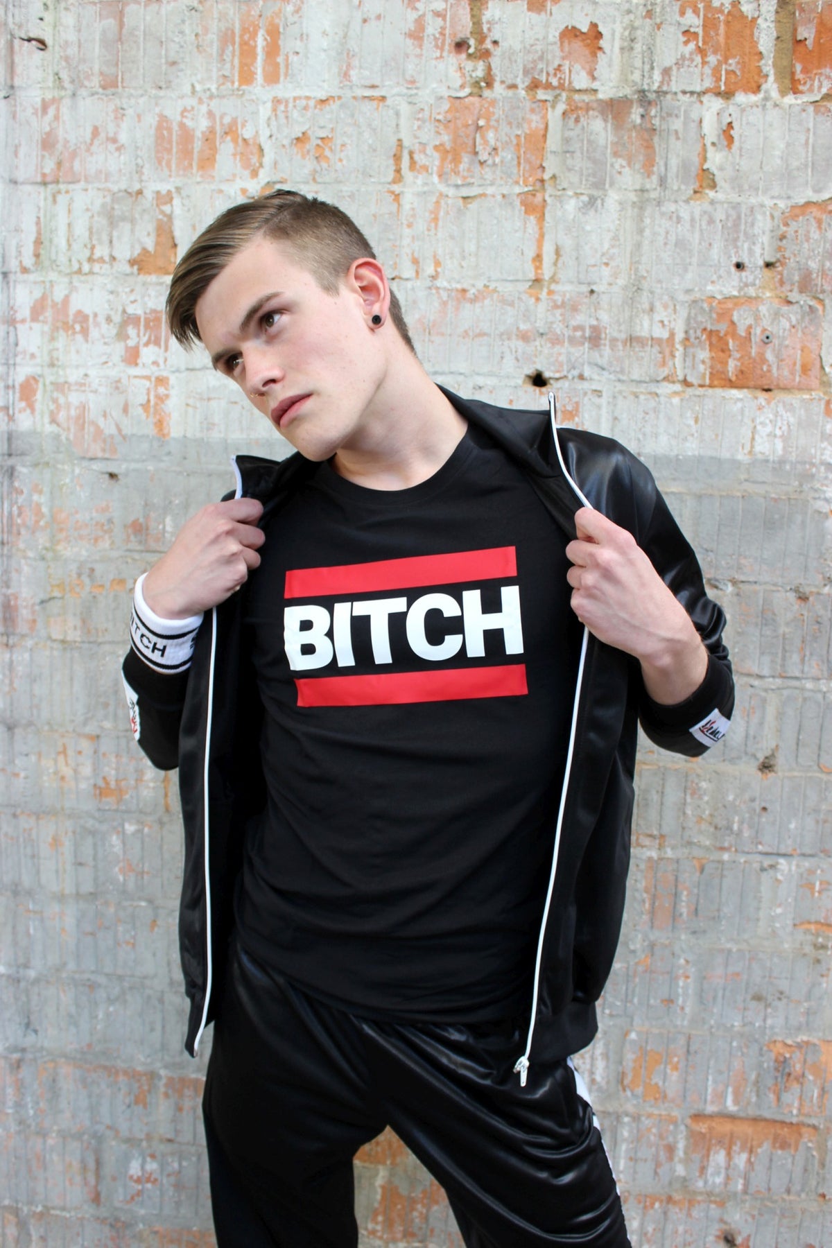 Young man holds his shiny sk8erboy jacket open, showing a black T-shirt with BITCH in uppercase and a red bar above and below.