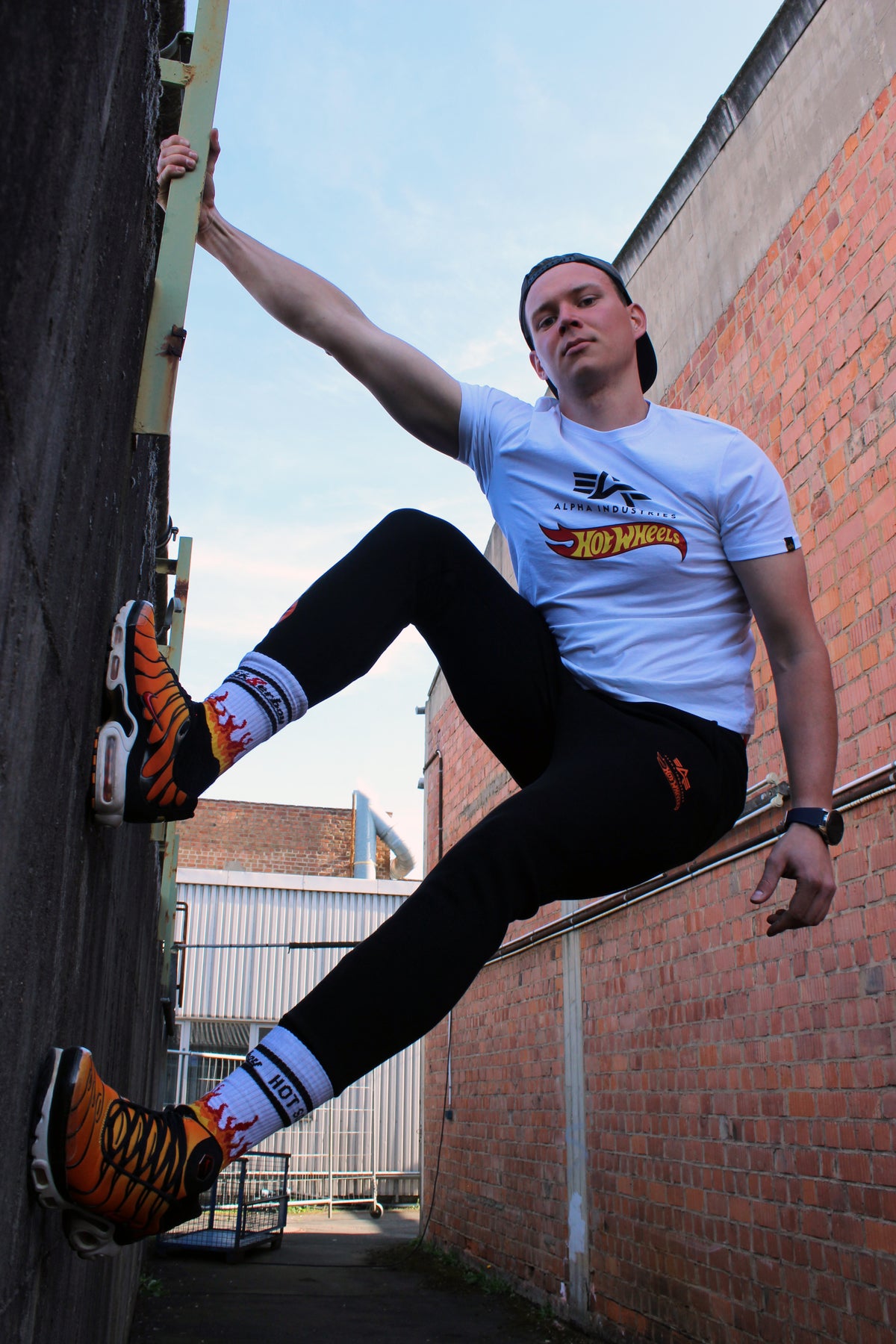 sk8erboy hot socks weisse socken mit schwarzem Schriftzug HOT SOCKS in grossen Buchstaben mit einer schwarzen Linie strich oben und unten ansonsten haben die socken ein flammen muster am Schaft und im fussbereich die flammen sind rot orange und gelb mit einer schwarzen Absetzung der boy Kerl traegt tiger nike tn tuned mit einem roten swosh haken er steht auf einer Leiter die an einem Feuerwehr auto steht mauer haengt