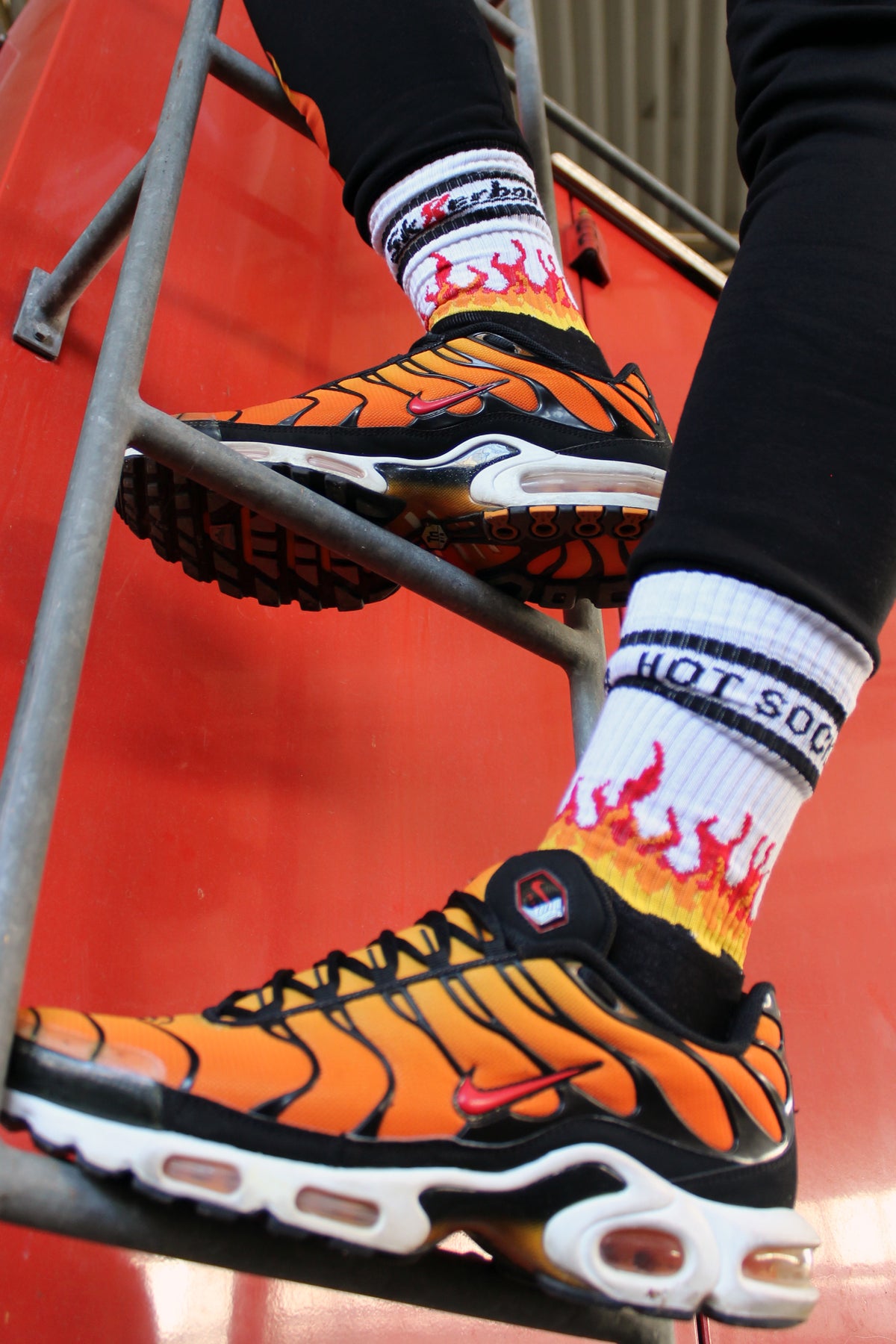 sk8erboy hot socks weisse socken mit schwarzem Schriftzug HOT SOCKS in grossen Buchstaben mit einer schwarzen Linie strich oben und unten ansonsten haben die socken ein flammen muster am Schaft und im fussbereich die flammen sind rot orange und gelb mit einer schwarzen Absetzung der boy Kerl traegt tiger nike tn tuned mit einem roten swosh haken er steht auf einer Leiter die an einem Feuerwehr auto steht