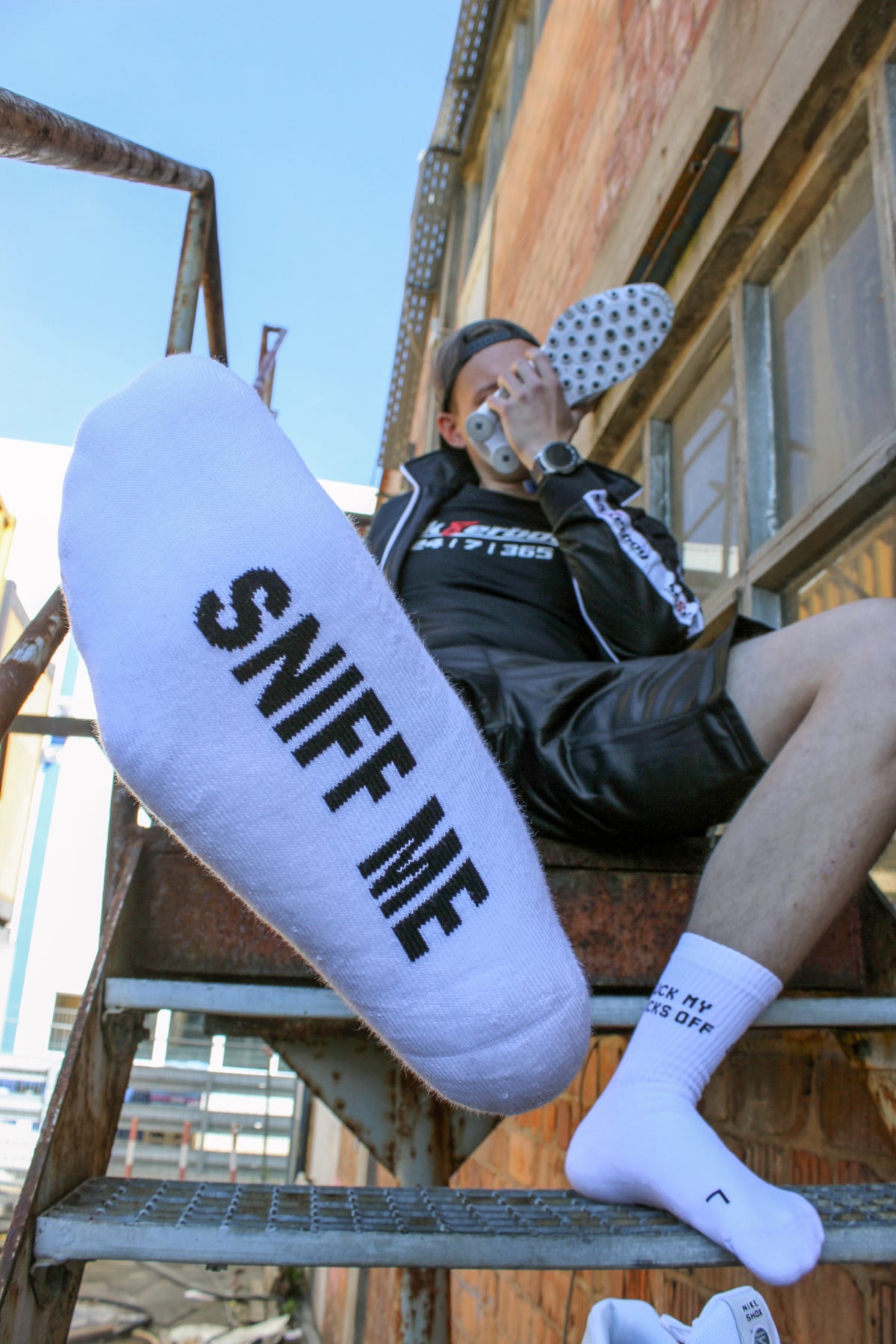 sk8erboy Sniff Me socks with bold black lettering on white socks; the boy sniffs his white leather Nike Shox. He wears black shiny sk8erboy shorts and jacket with sk8erboy embroidery, plus a black cotton T-shirt with 24 7 365 in white and a red 8.
