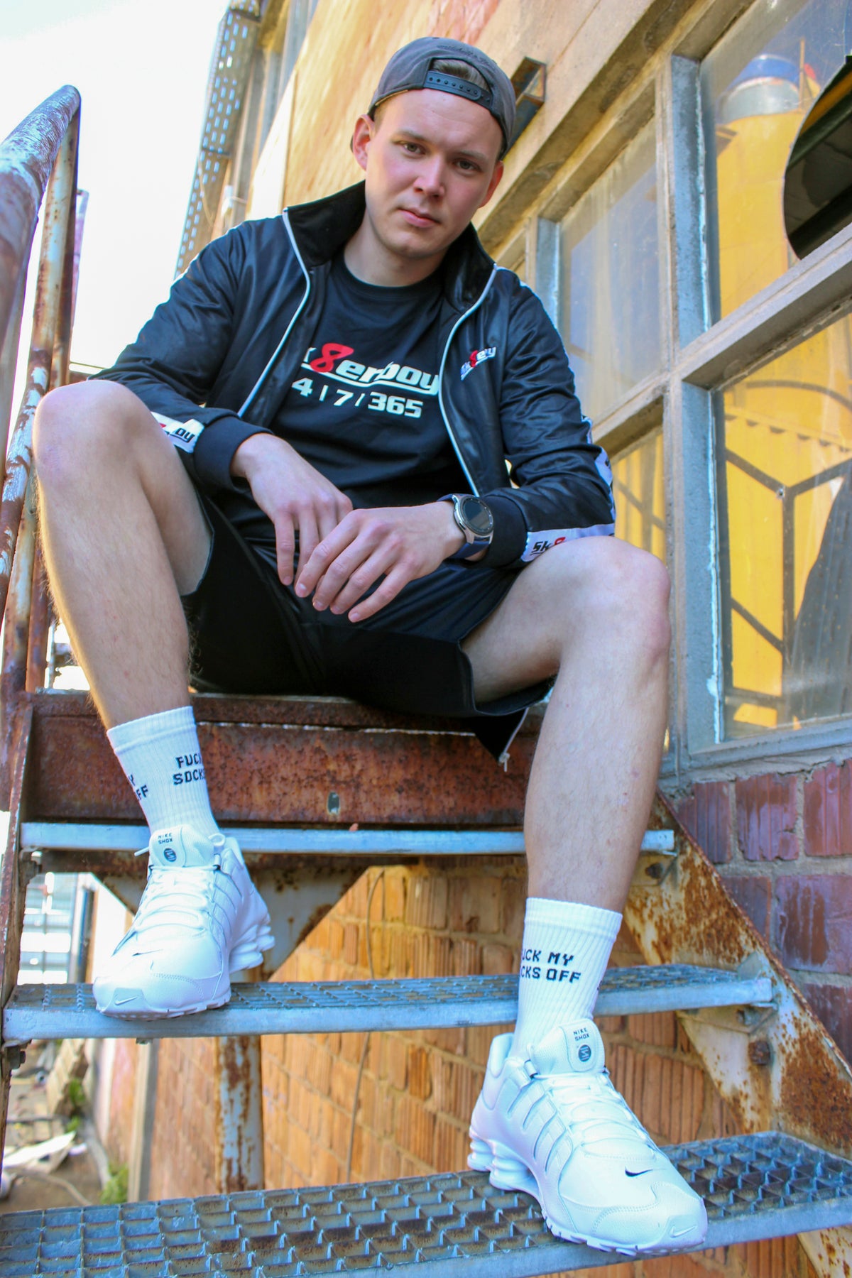 sk8erboy sniff me socks, white socks with bold black print. The boy sniffs hard on his white leather Nike Shox, wearing sk8erboy shiny black shorts and a glossy black sk8erboy jacket with chest embroidery. T-shirt reads 24/7/365 and red 8.