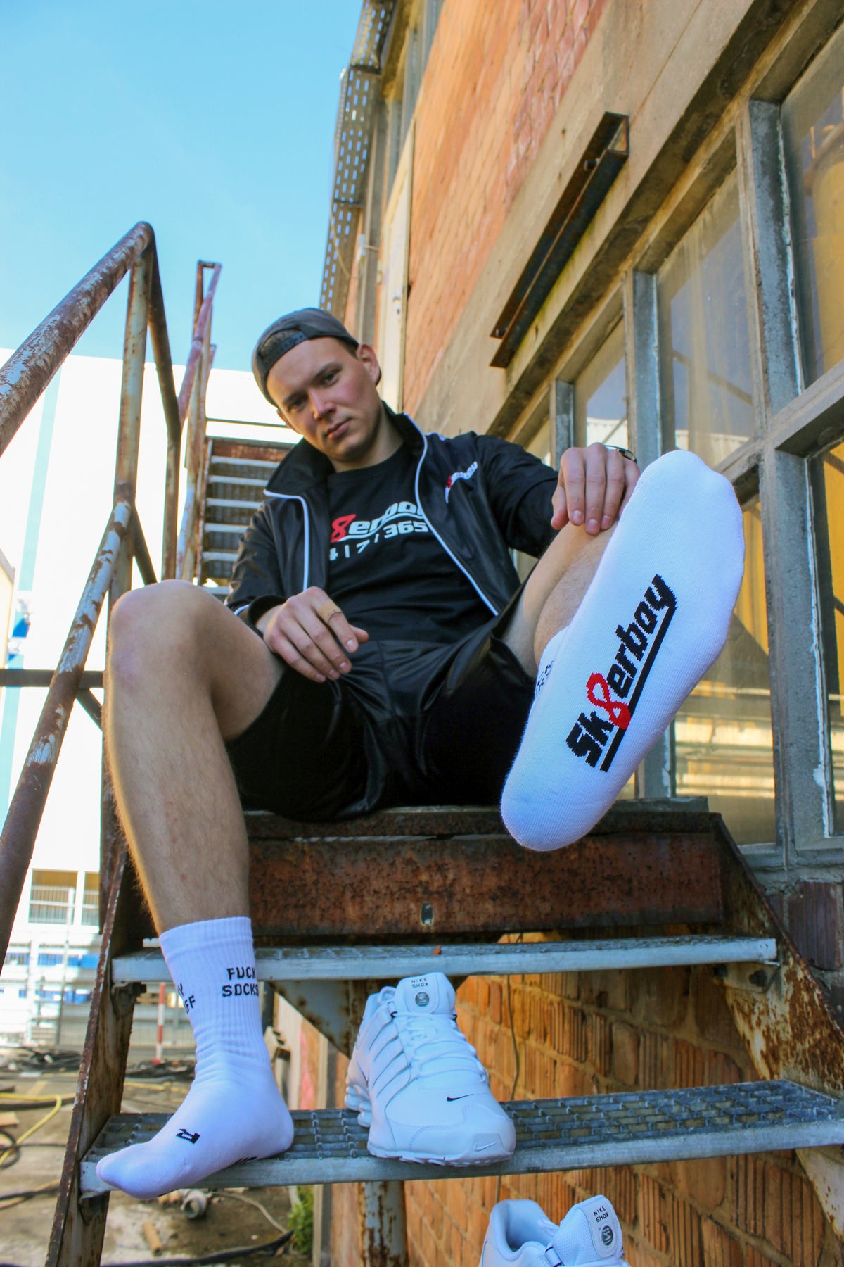 sk8erboy sniff me socks with bold black lettering on white socks; the boy sniffs his white leather Nike Shox, wearing black glossy sk8erboy shiny shorts and a shiny black sk8erboy jacket with chest embroidery, plus a T-shirt with 24 7 365