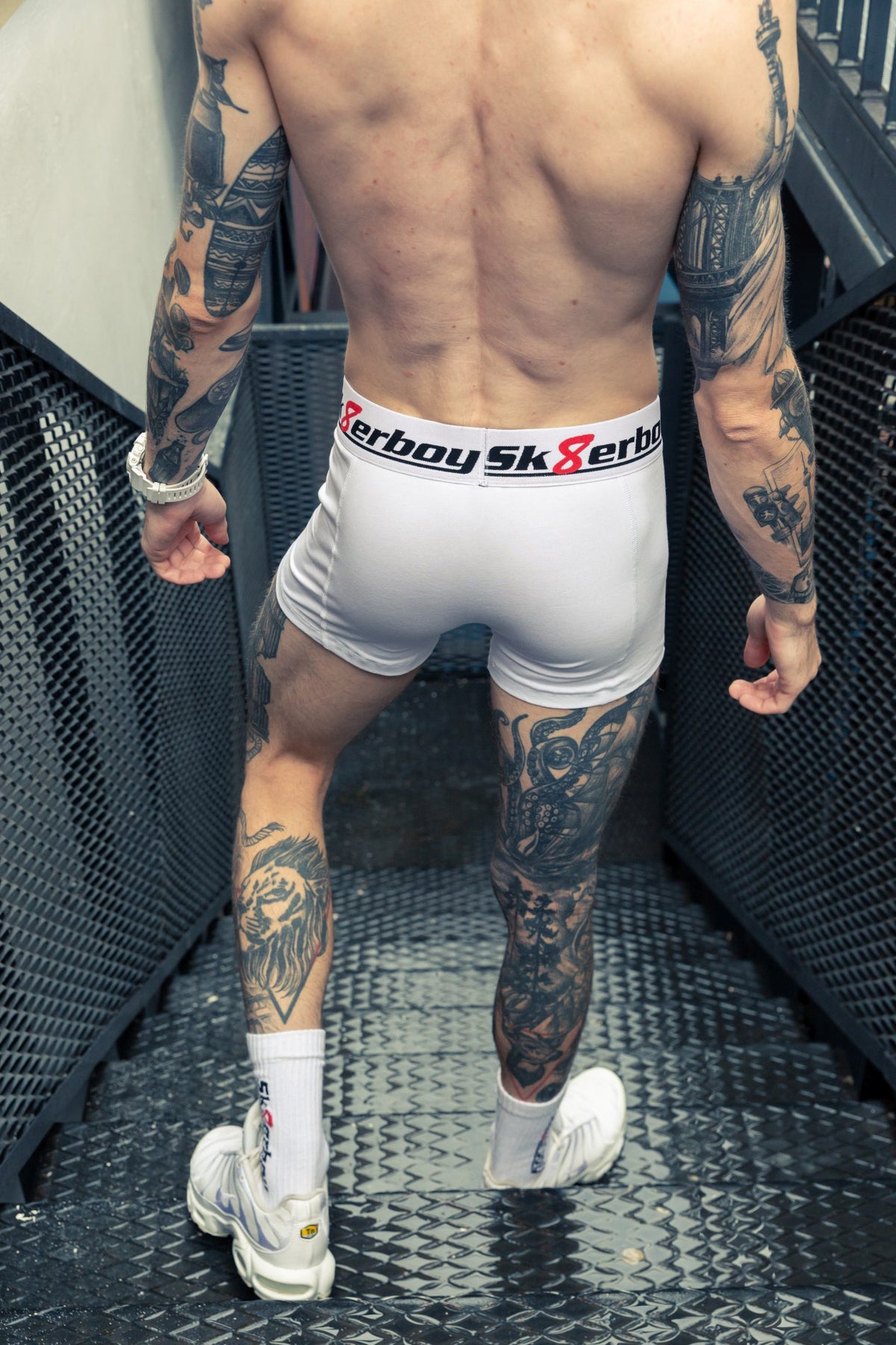 Athletic man from behind showing his firm butt, wearing white SK8ERBOY boxers with a large stylish logo waistband and matching SK8ERBOY crew socks in Nike TN sneakers on an industrial staircase