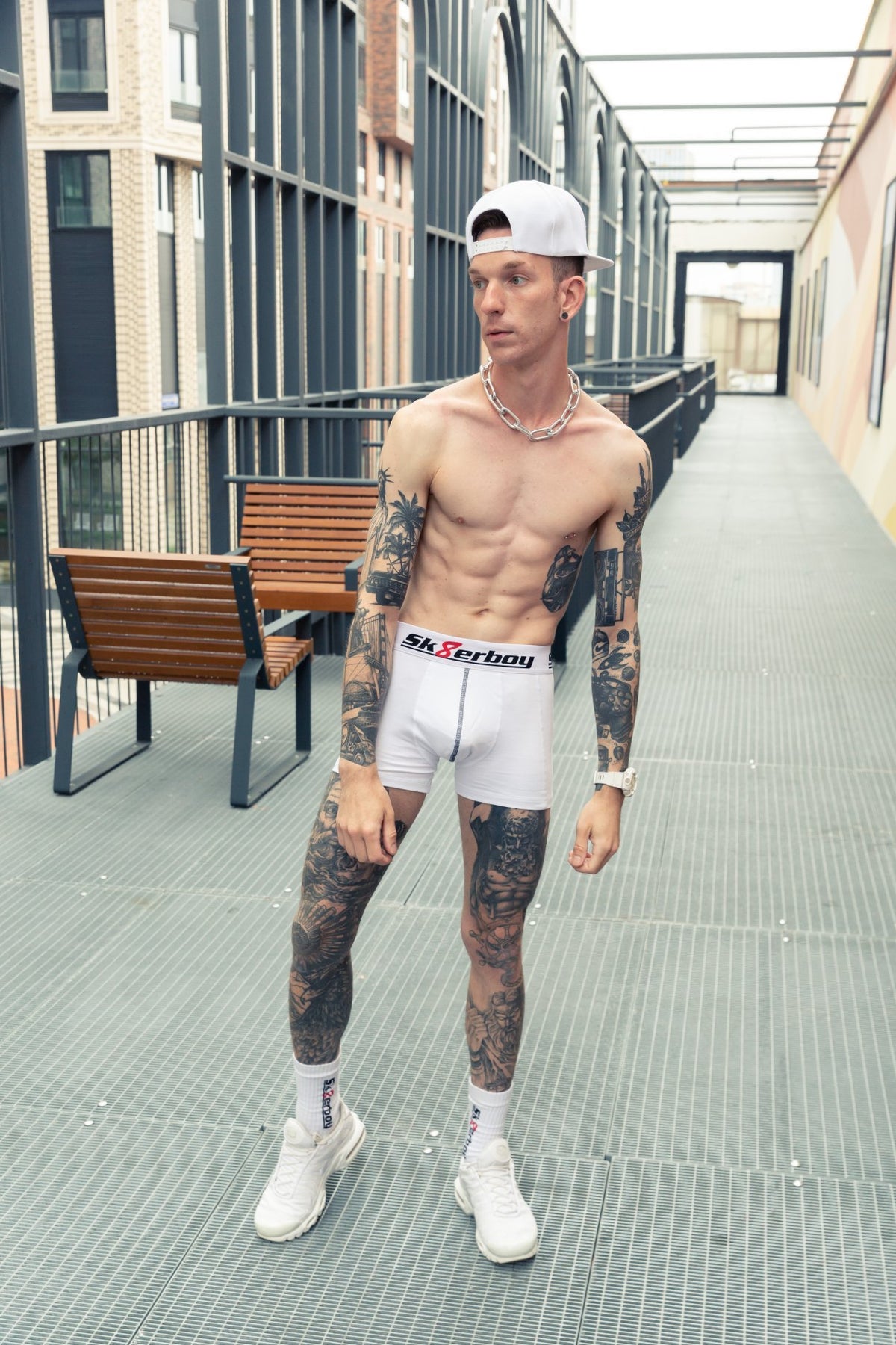 Fit young man with bare chest and tattoos on legs and arms wearing white SK8ERBOY boxer shorts, matching white crew socks, and Nike TN sneakers, with a cap and a steel chain necklace.