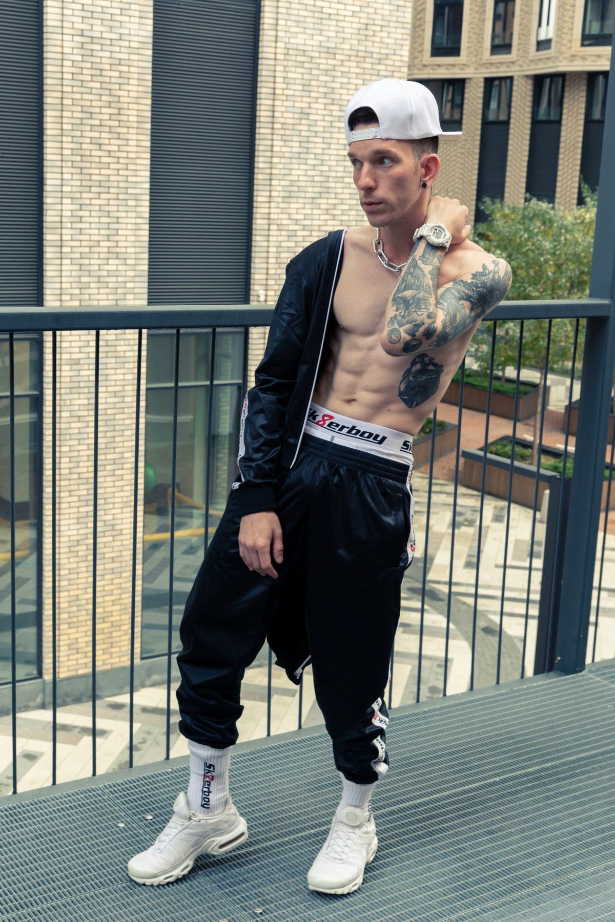 Athletic young man wearing a shiny black tracksuit and matching crew socks with white boxershorts by sk8erboy in a city on a terrace, with white Nike TN sneakers and a cap