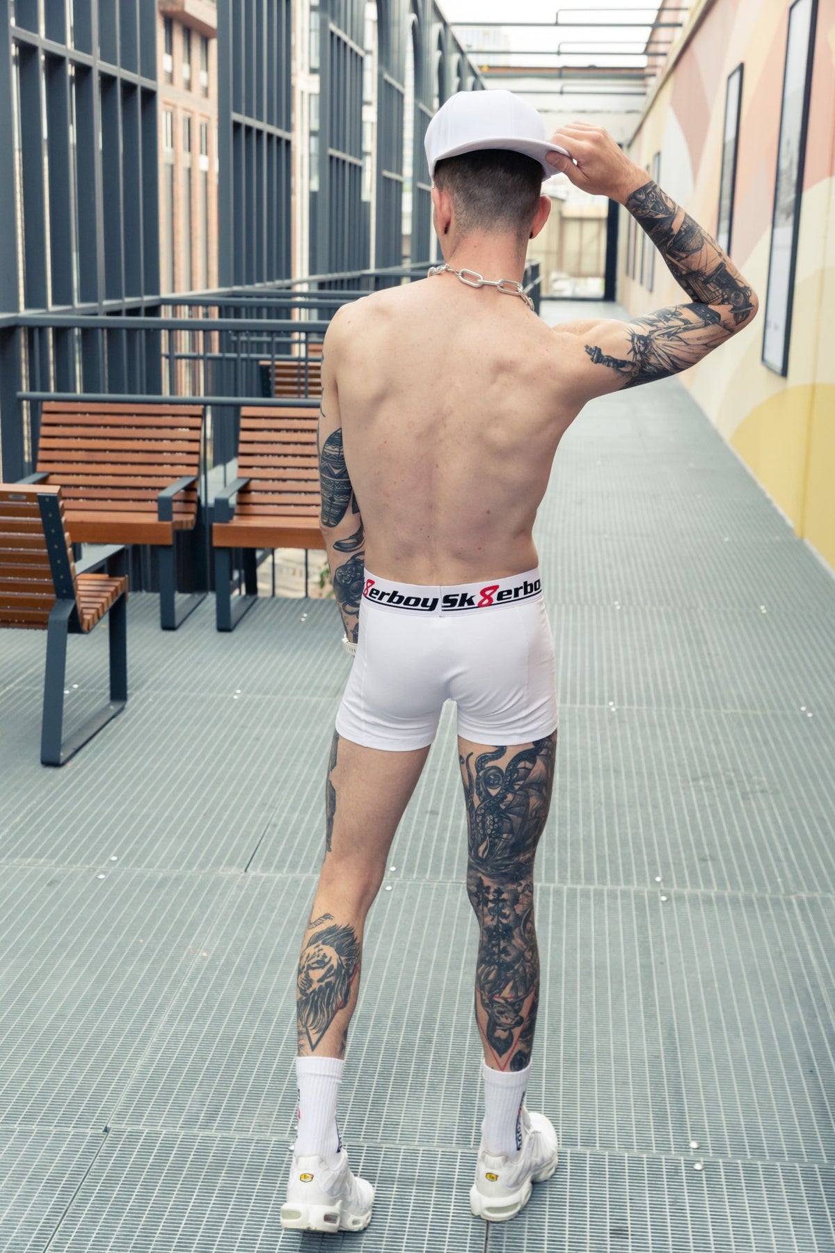 athletic young man seen from behind, shirtless, wearing white Sk8erboy boxer shorts with logo on the waistband and matching crew socks in white Nike TN sneakers with cap