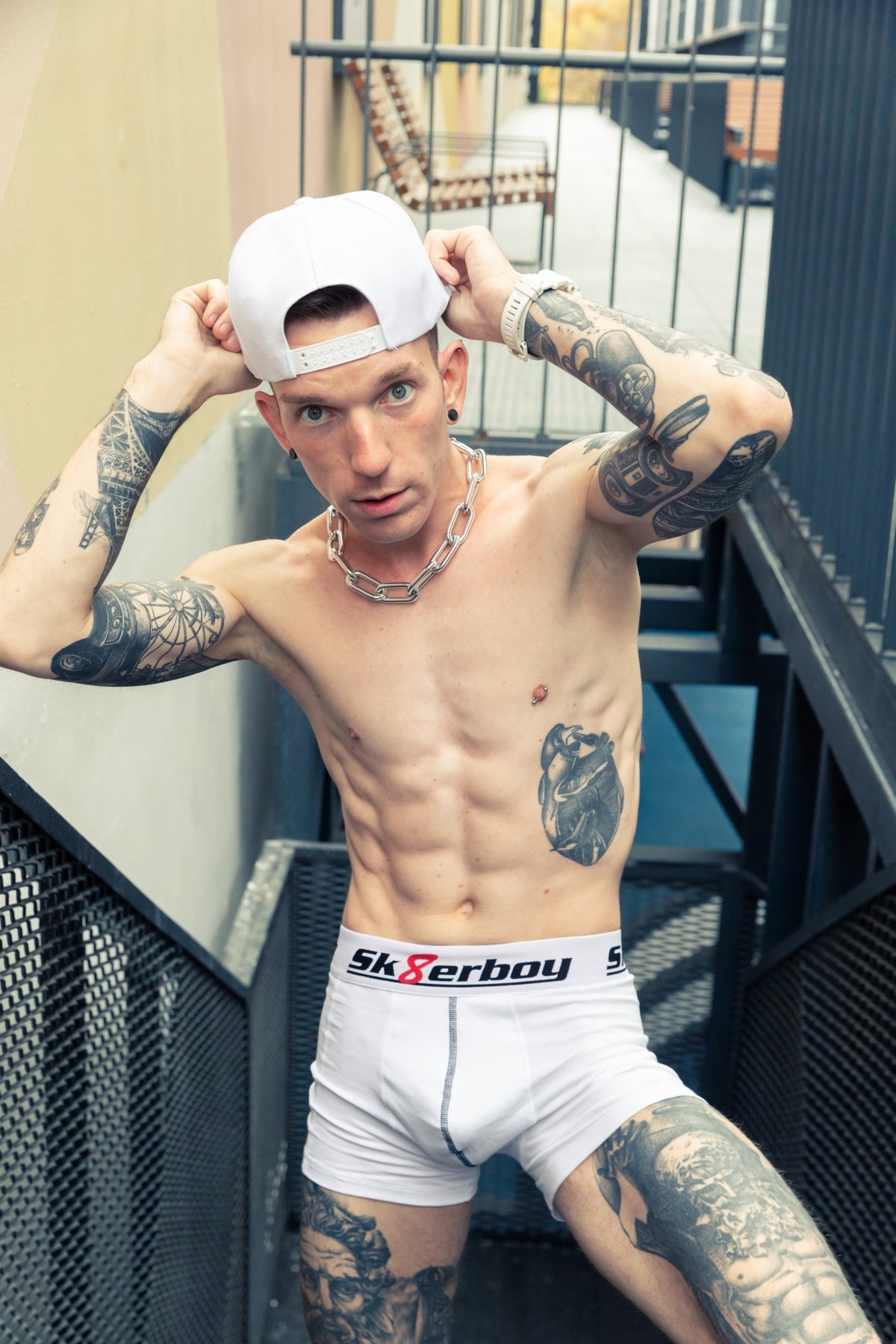 Athletic upper body of a young man from the front, wearing white boxershorts by sk8erboy and a thick chain necklace, with a cap on a staircase