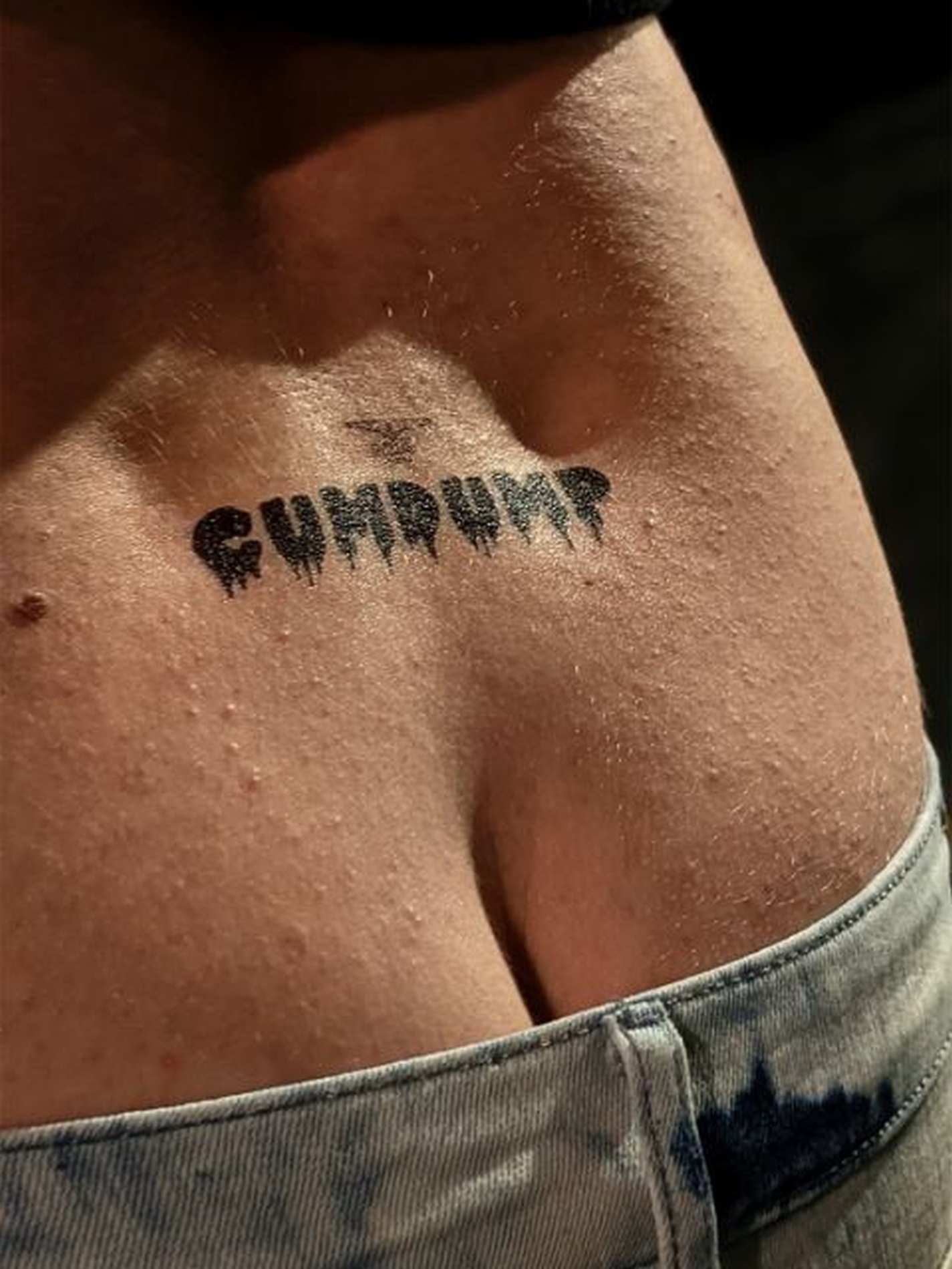 ansicht eines männerrückens vor einer dunklen wand der ein temporäres tattoo von mister b über dem po und der aufschrift cumdump in schwarz und bitch in rot trägt.
