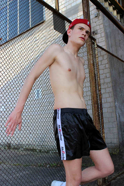 junge mit roter kappe an einem zaun und freiem oberkoerper mit sneaker von nike traegt eine schwarze shiny boxershort unterwaesche von sk8erboy mit banner seitlich